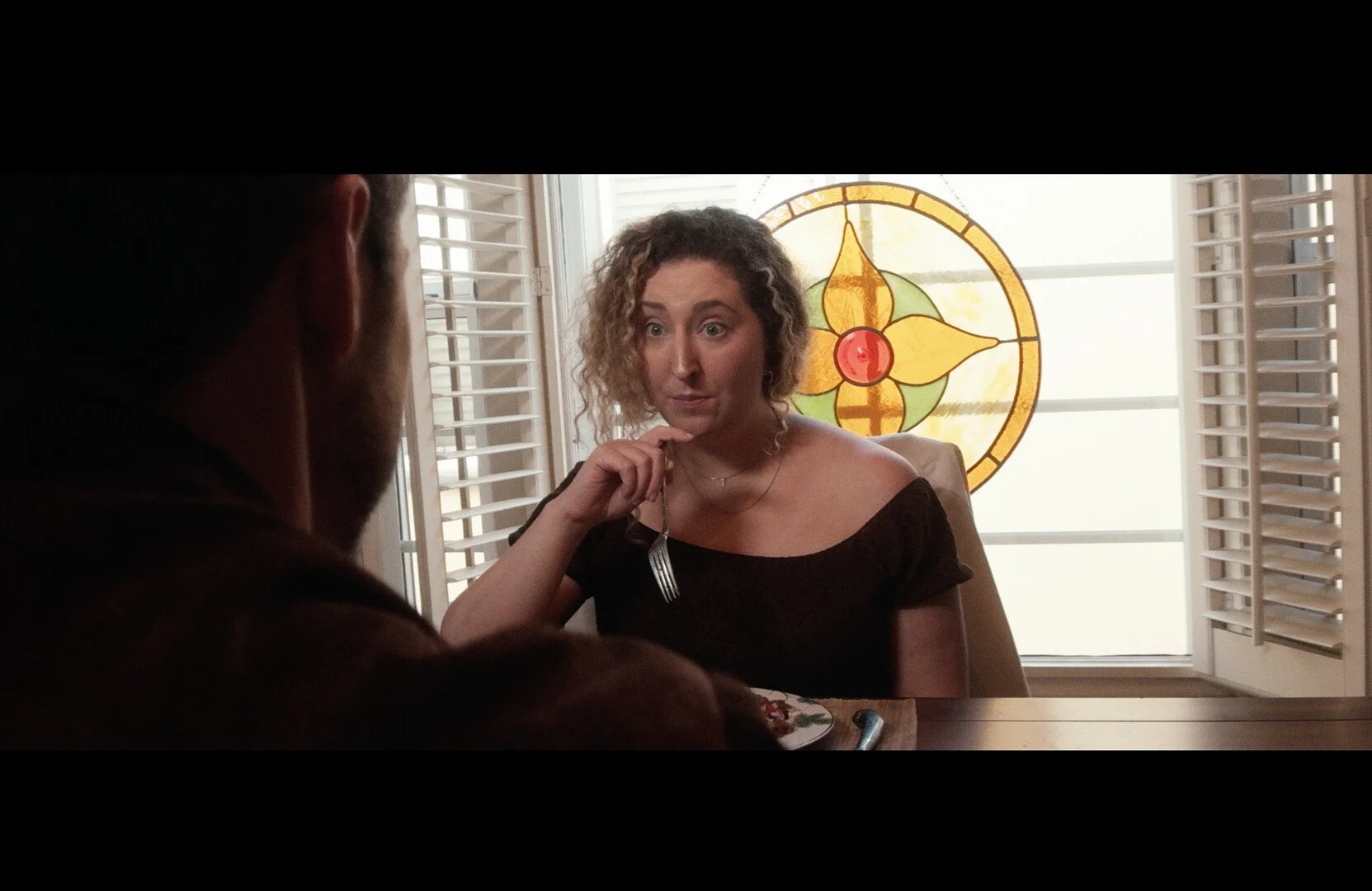 A woman sitting at a dining table with a man in a dimly lit room, with a large colorful stained glass window featuring a floral design in the background.