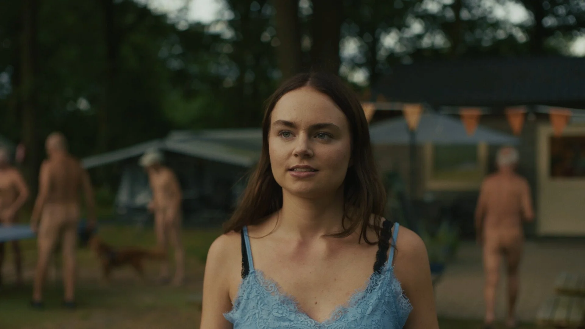 A woman with brown hair in a blue lace dress standing outdoors, with several people in the background in a park or garden setting.