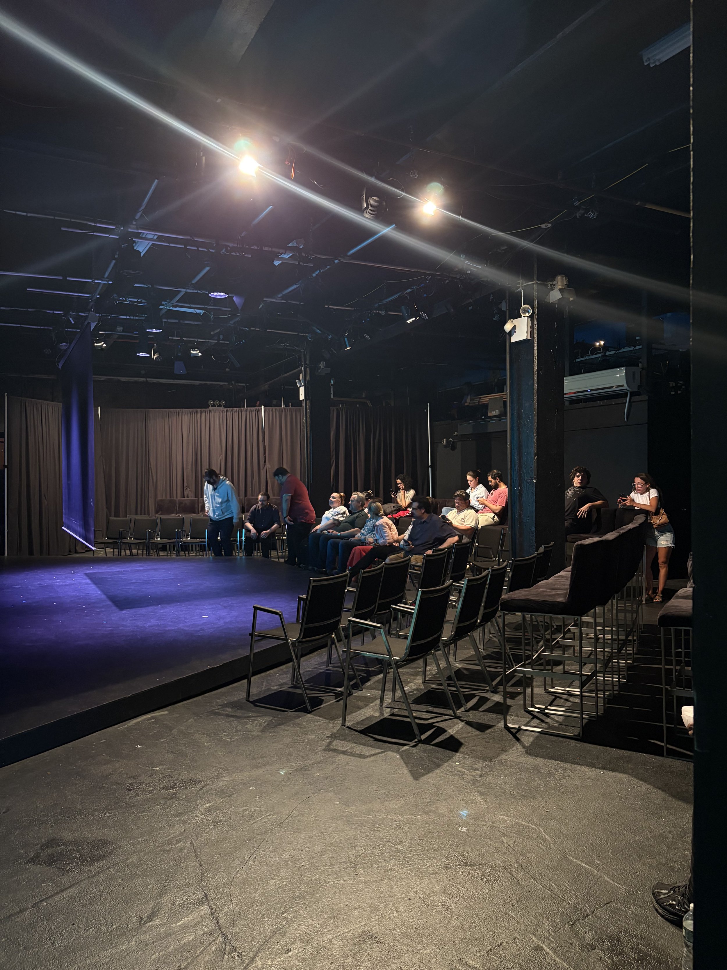 People seated and standing in a dimly lit theater or comedy club with chairs and a stage, some waiting or socializing.