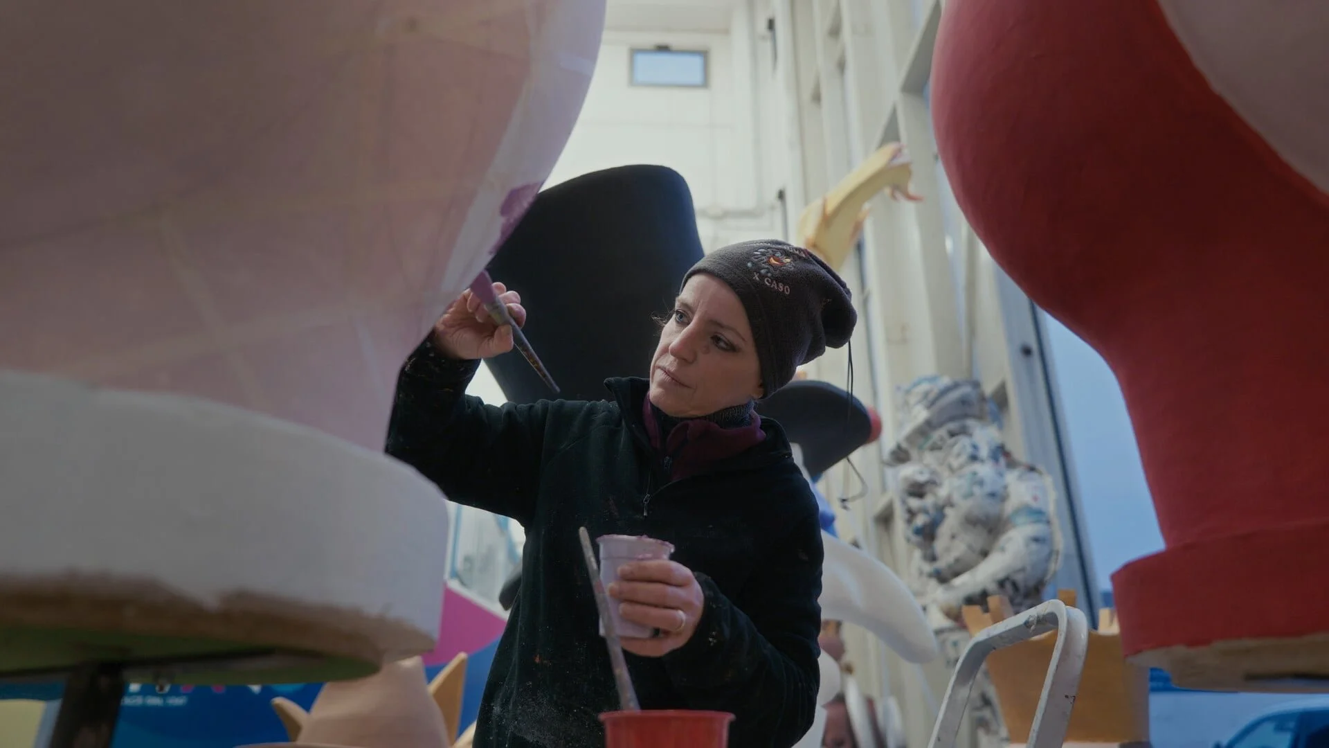 A woman wearing a black beanie and dark clothing decorates ceramic vases in a ceramics studio, with large pottery pieces around her.