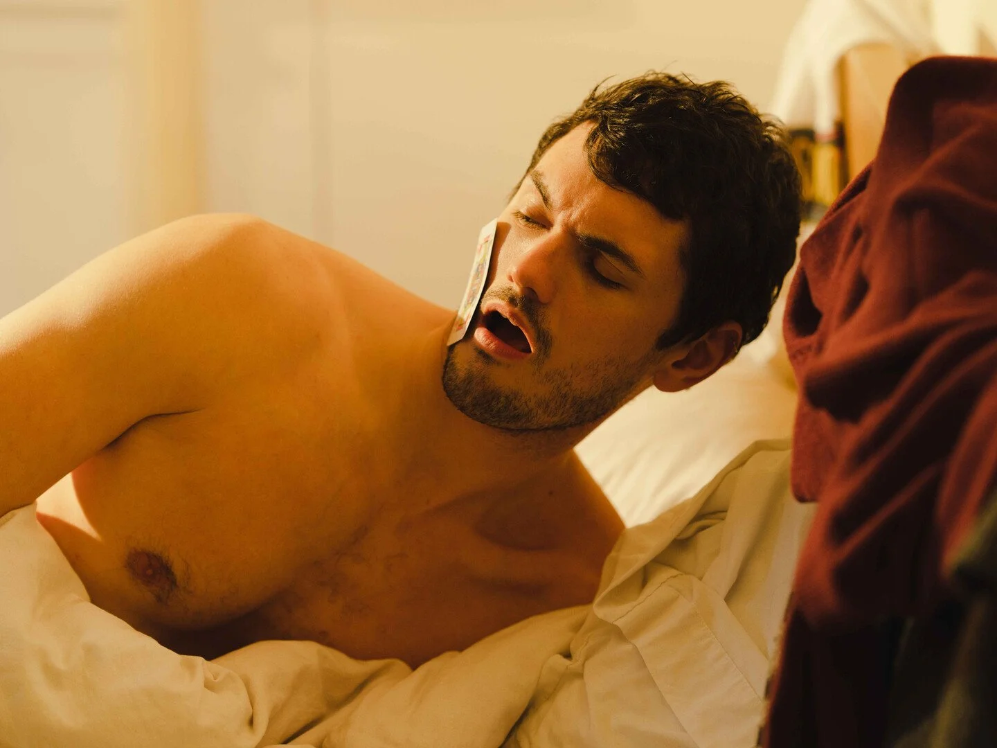 A shirtless man with a bandage on his face lying in bed with eyes closed.