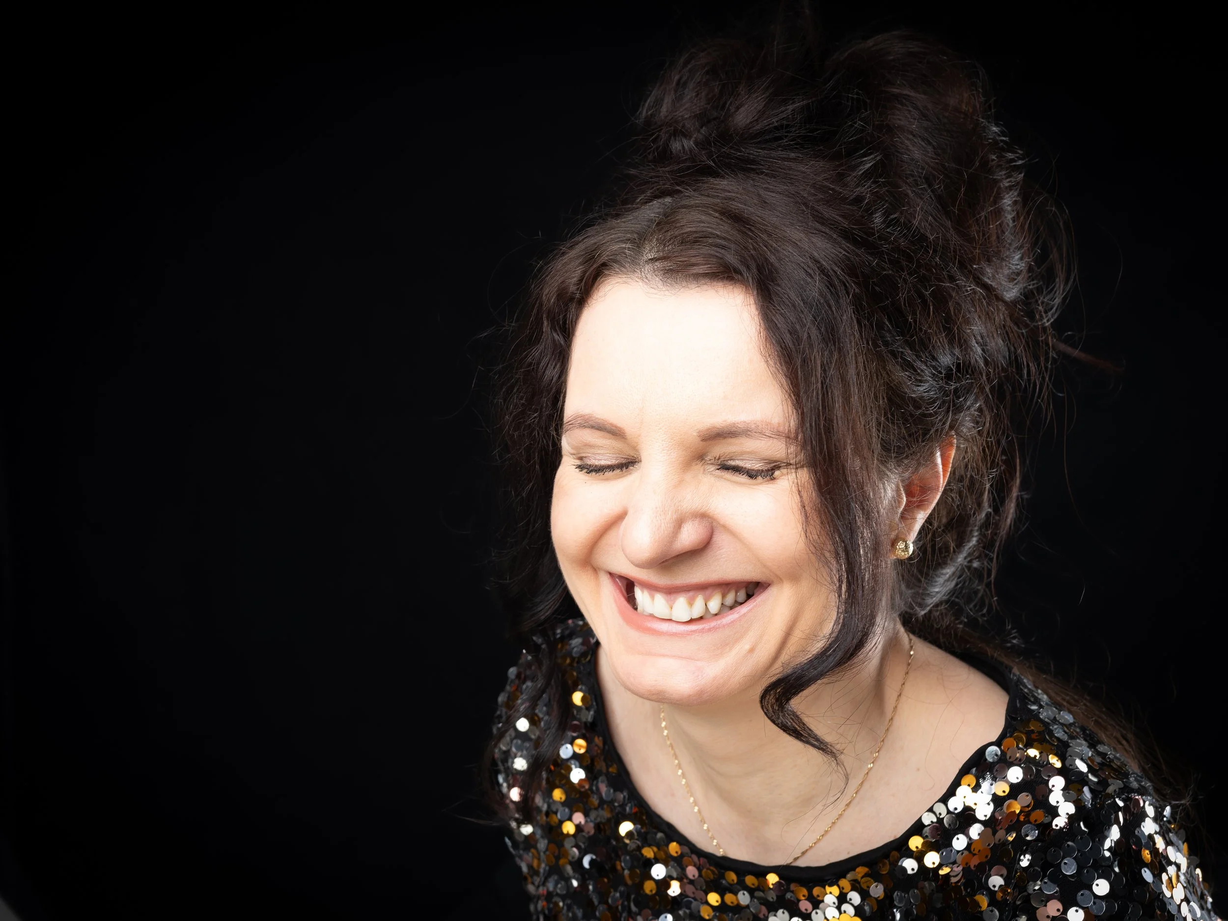 Smiling woman with dark, curly hair styled up, wearing a black sequin top and gold jewelry, against a black background.