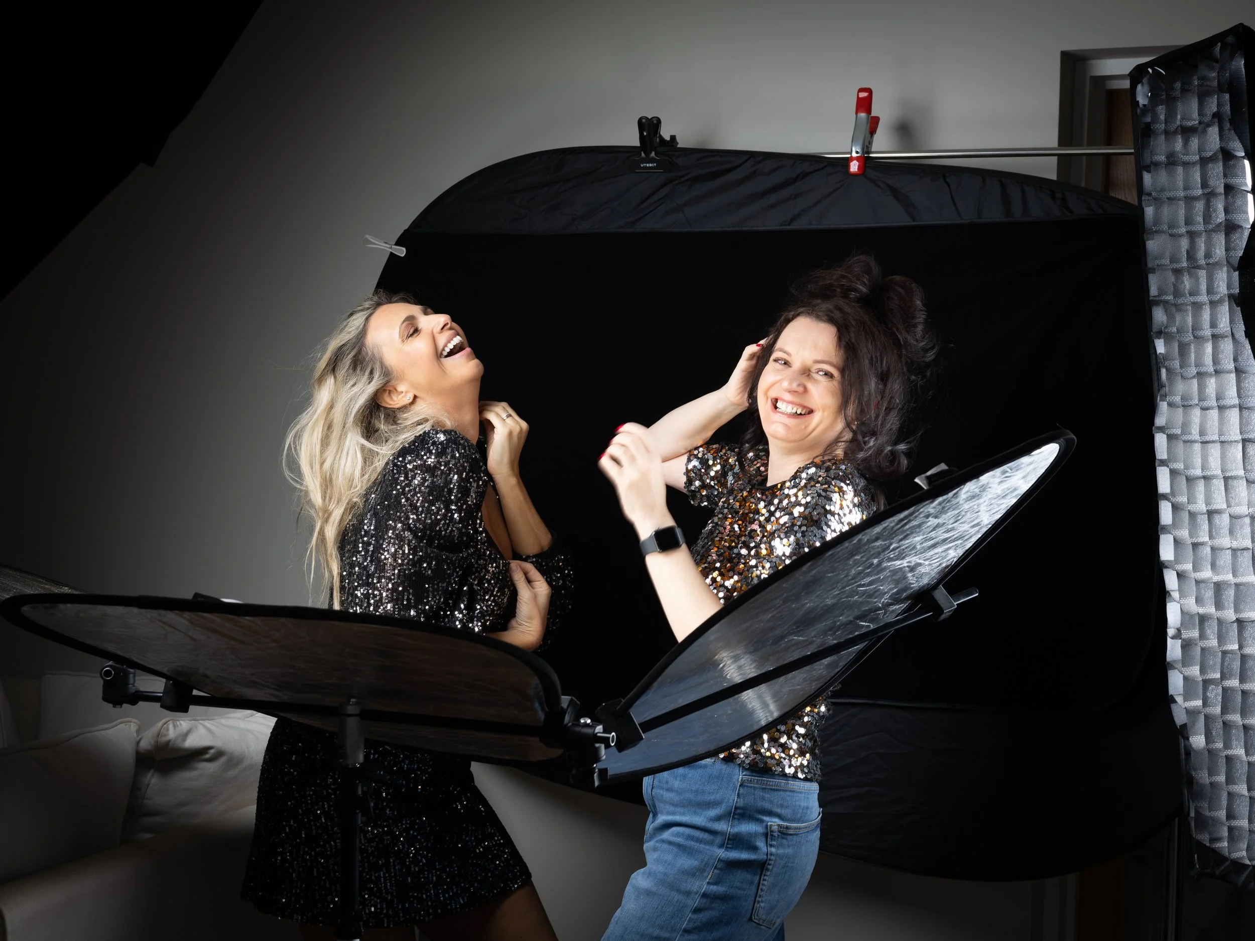 Two women with happy expressions standing next to a grand piano, one touching the other's head, in a photo studio with a black backdrop and a gray curtain on the right.