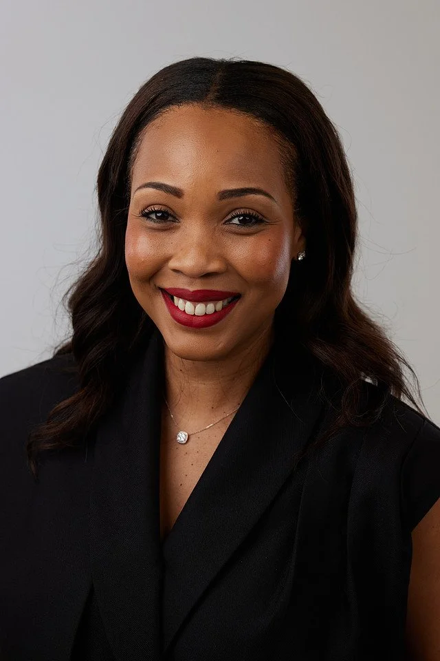 Professional woman with shoulder-length brown hair, red lipstick, wearing pearl and Chanel jewelry, black top, and houndstooth blazer against a white background.