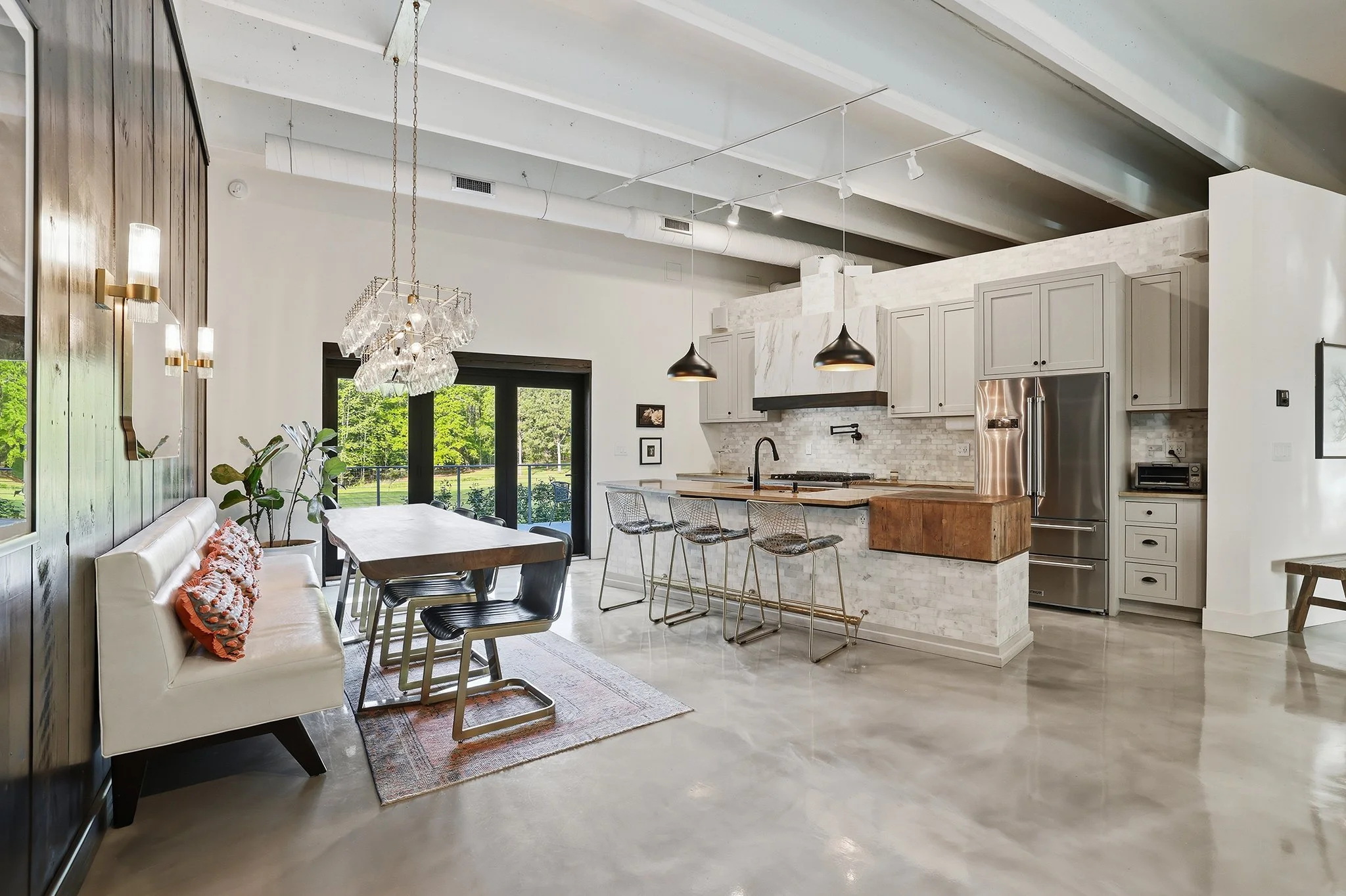 Modern kitchen and dining area with a white kitchen, marble backsplash, and a large island with barstools. There's a dining table with chairs, a white bench with pillows, and sliding glass doors leading outside. The space features high ceilings, pendant lights, and large windows.
