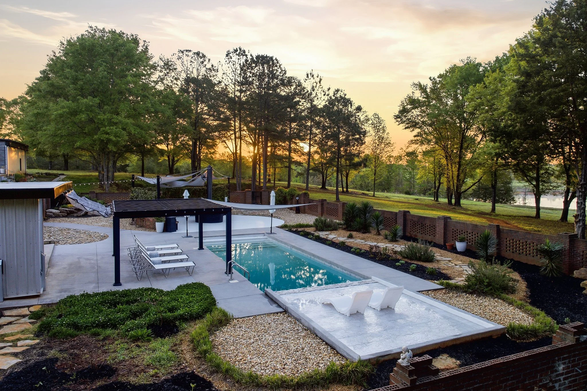 A backyard with a swimming pool and lounge chairs during sunset, surrounded by trees, plants, and a brick wall fence.