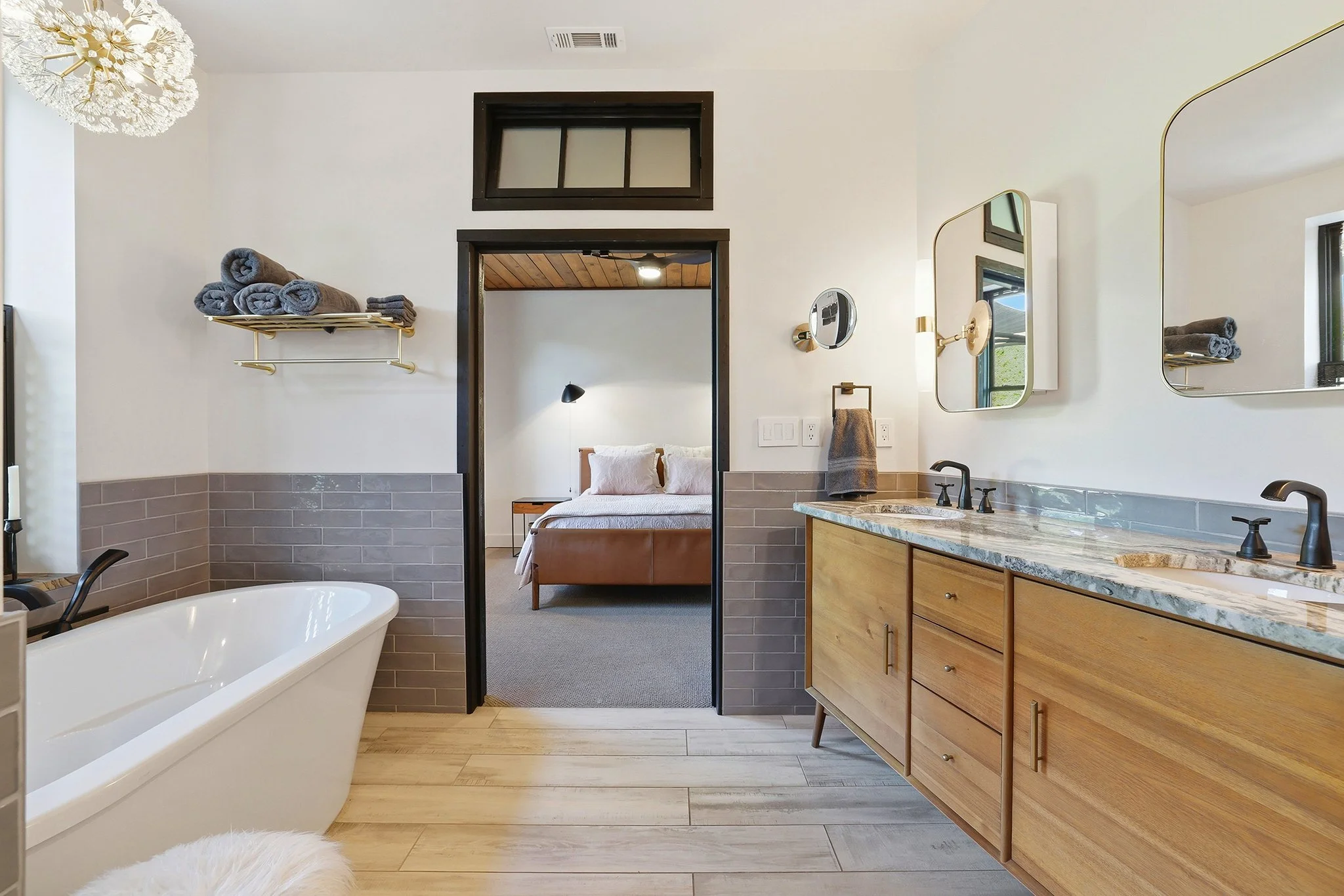 A modern bathroom with a freestanding bathtub, a wooden vanity with a marble countertop, two mirrors, and gray towels on a shelf. The room opens into a bedroom with a bed and pendant lamp visible through the doorway.
