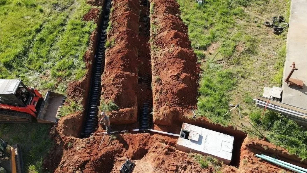 Construction site with a trench being dug, black corrugated pipes laid inside, and a small construction vehicle at the bottom left. Green grass surrounds the trench, and construction materials are on the right side.