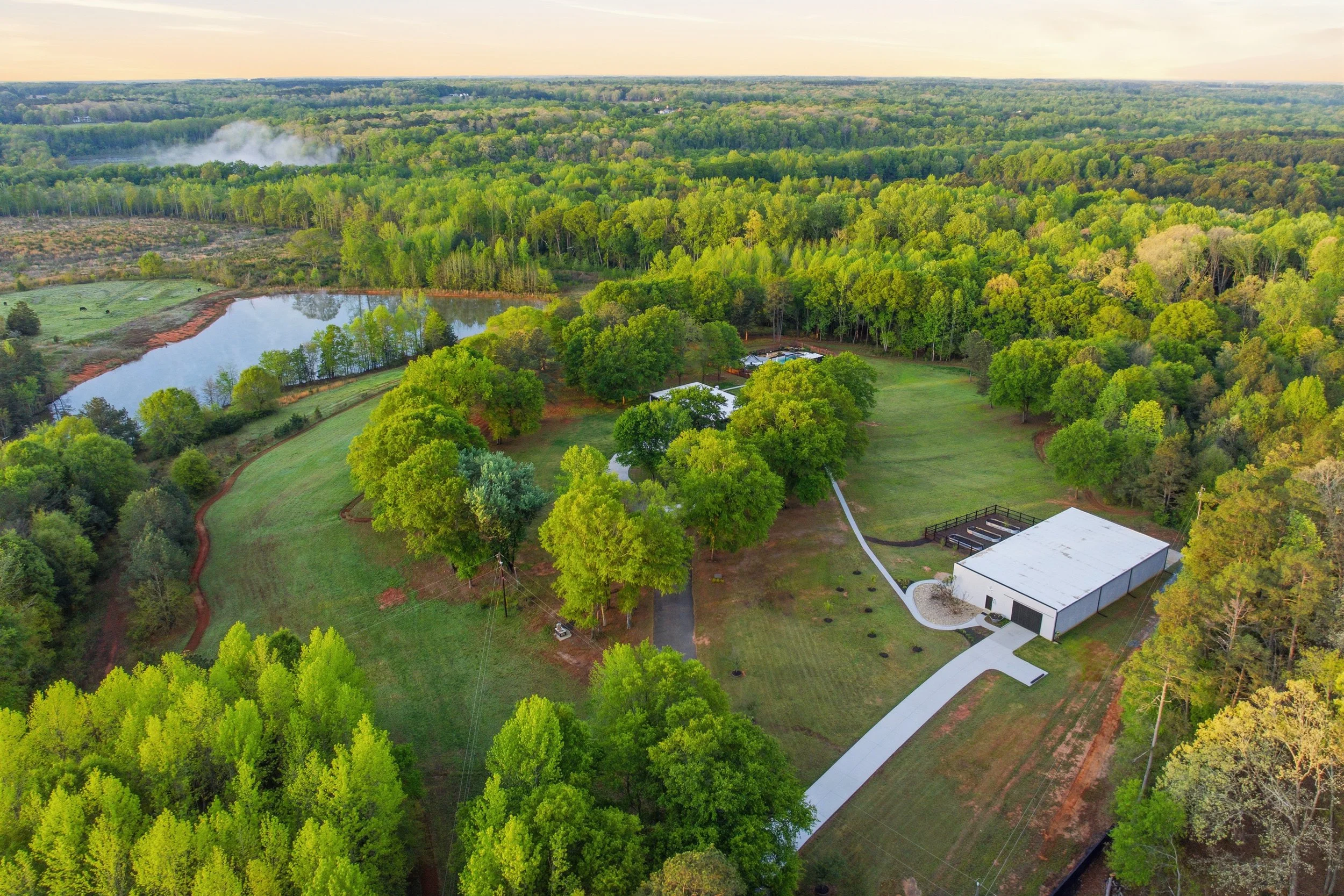 Aerial view of a rural property surrounded by lush green trees, with a pond on the left and a large white building with a curved driveway on the right.