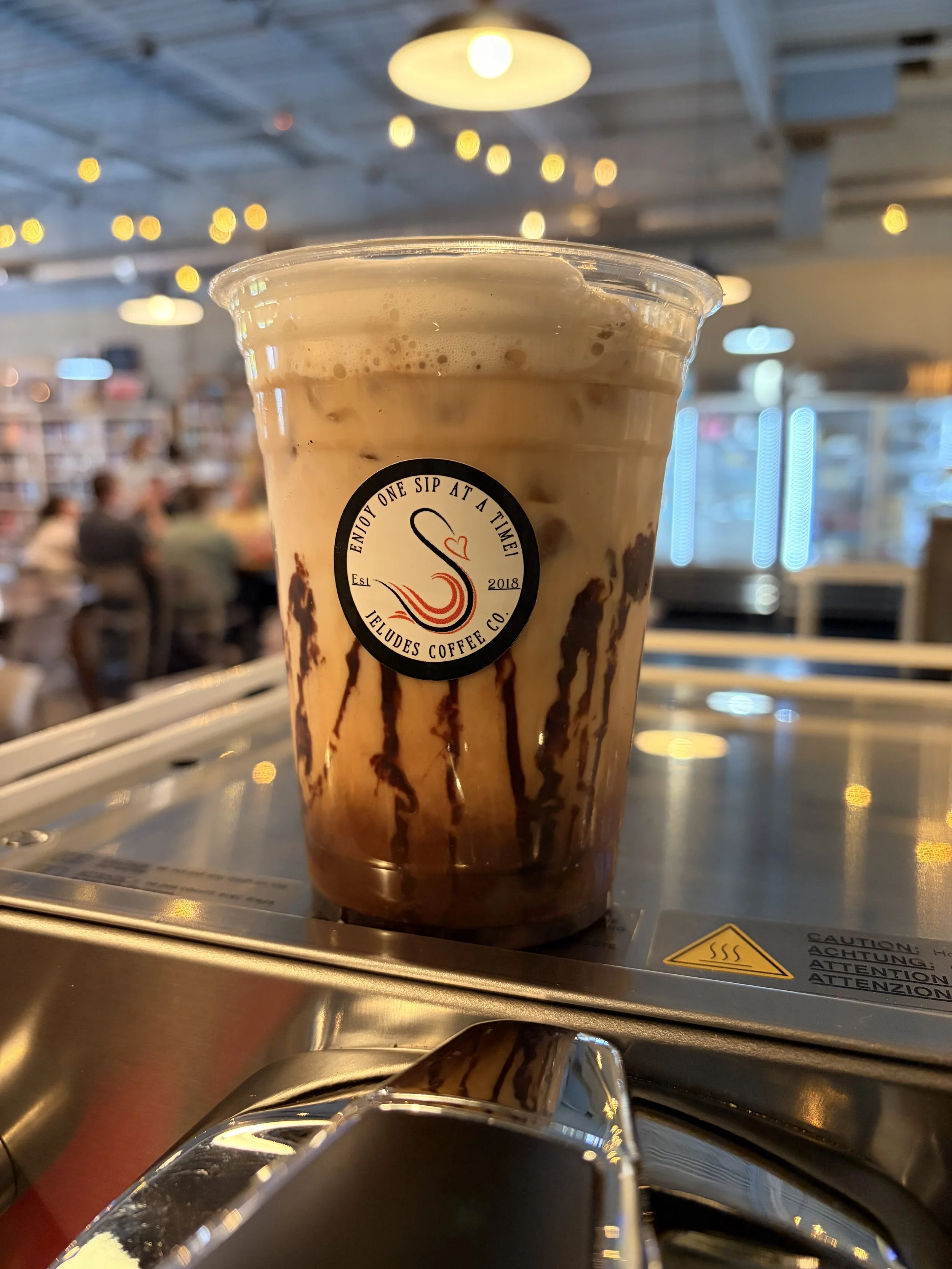 A plastic cup with iced coffee and chocolate syrup, with a logo saying 'Enjoy One Sip at a Time JeLudes Coffee Co.' In a coffee shop, with blurred customers in the background.