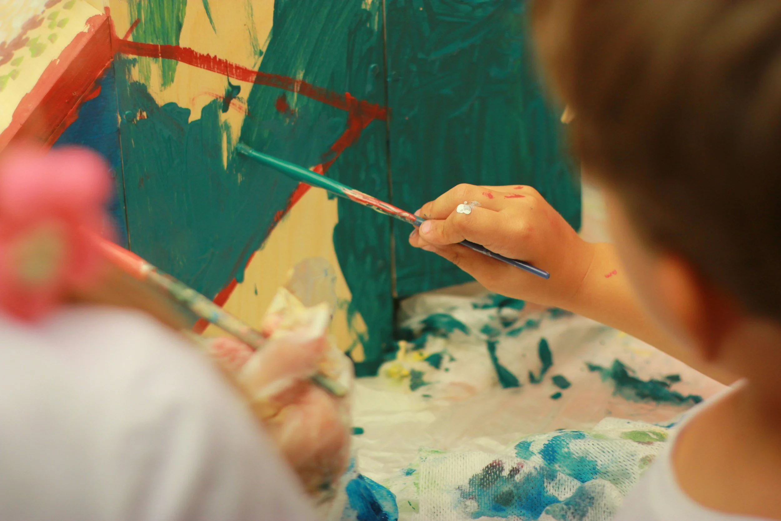 Close-up of a person's hands holding a paintbrush, painting on a multicolored canvas with green, yellow, red, and blue sections. The person's hands and the painting are expressive of artistic activity.