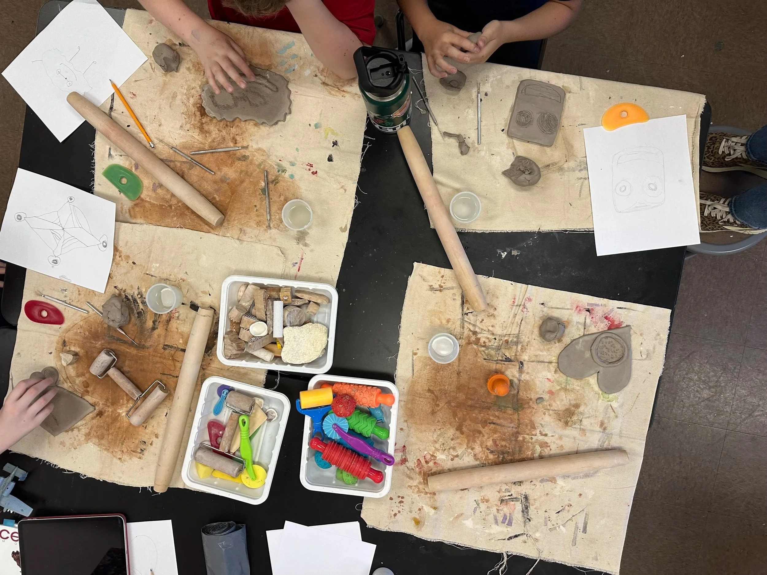 Children engaging in a pottery or sculpture class, working with clay and various tools on a table covered with a cloth. There are sketch drawings of possible clay designs and containers of small clay pieces and modeling tools.