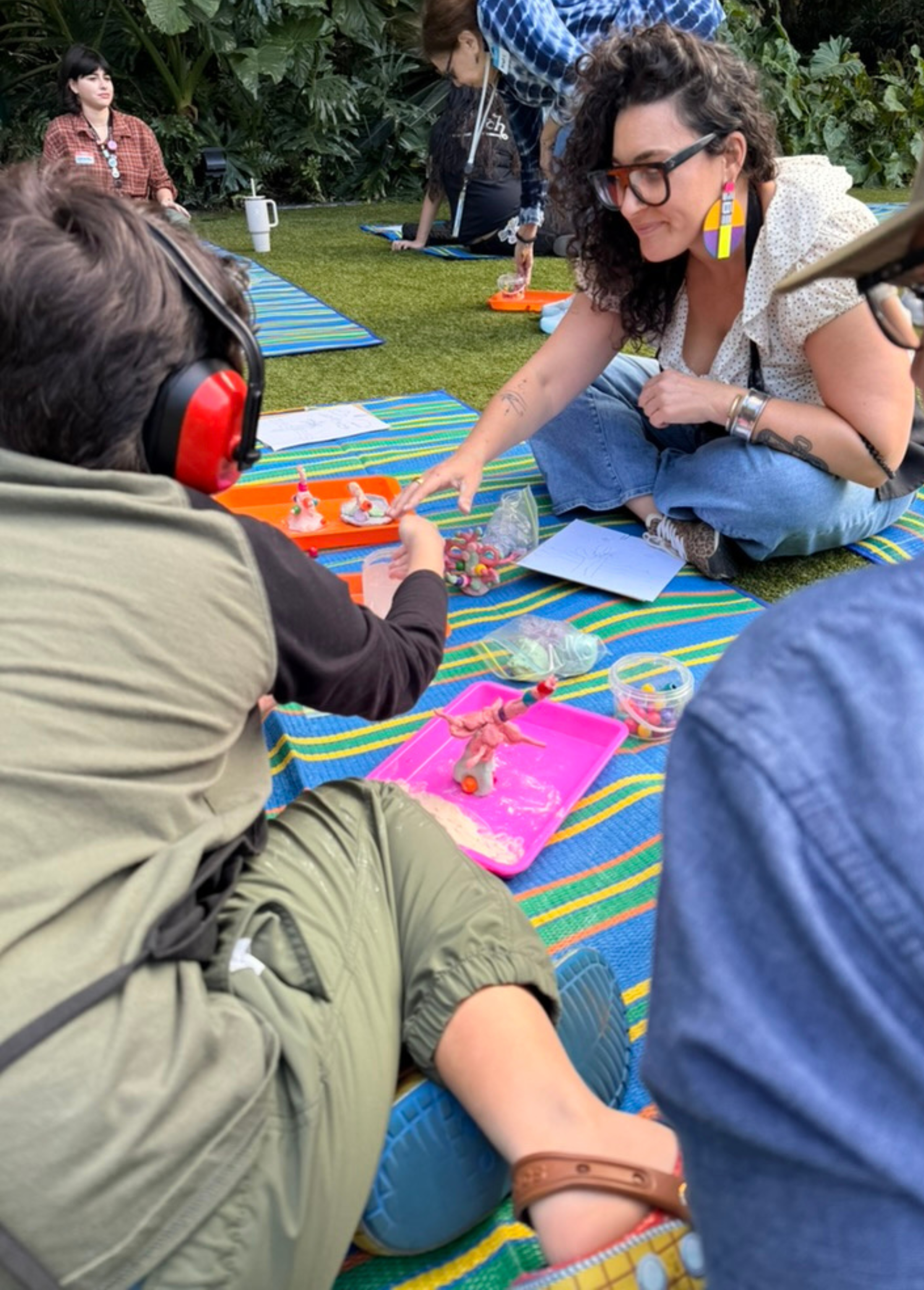 A woman with curly hair, glasses, and colorful earrings, sitting cross-legged on a striped mat, interacts with a child who is wearing large red ear protection and holding small figurines. The scene appears to be outdoors with green plants in the background, and other people are sitting or kneeling on similar mats, engaged in activities at a table with toys and papers.