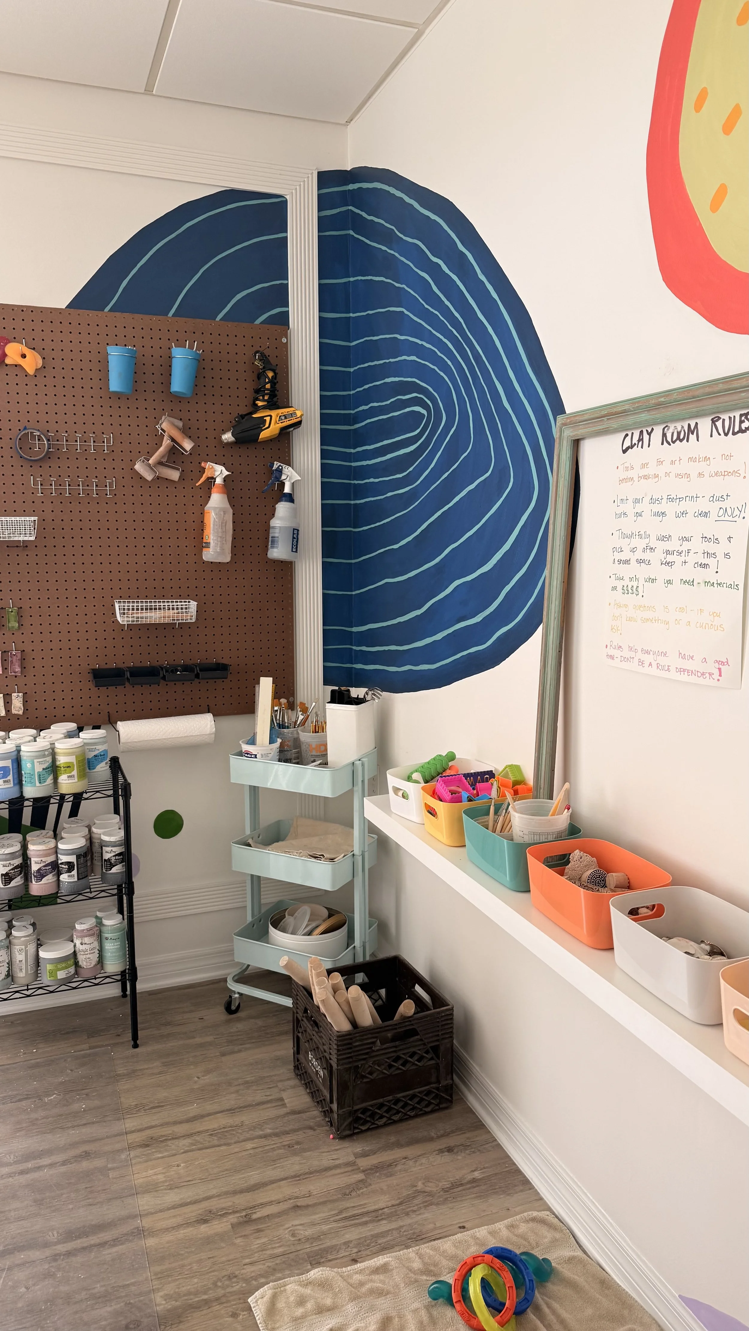 An art therapy or creative workspace with a pegboard holding supplies like glue guns and sprays. There's a white rolling cart and various containers of craft materials, paint, and tools. The wall has a blue spiral mural and a whiteboard with rules for a clay room. Colorful rings and toys are on the floor.