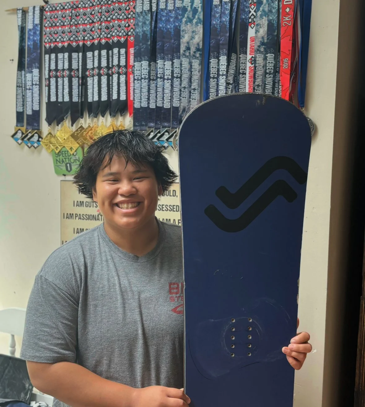 A smiling young person holding a blue skateboard with a black logo, standing in front of a wall decorated with racing medals and motivational posters.