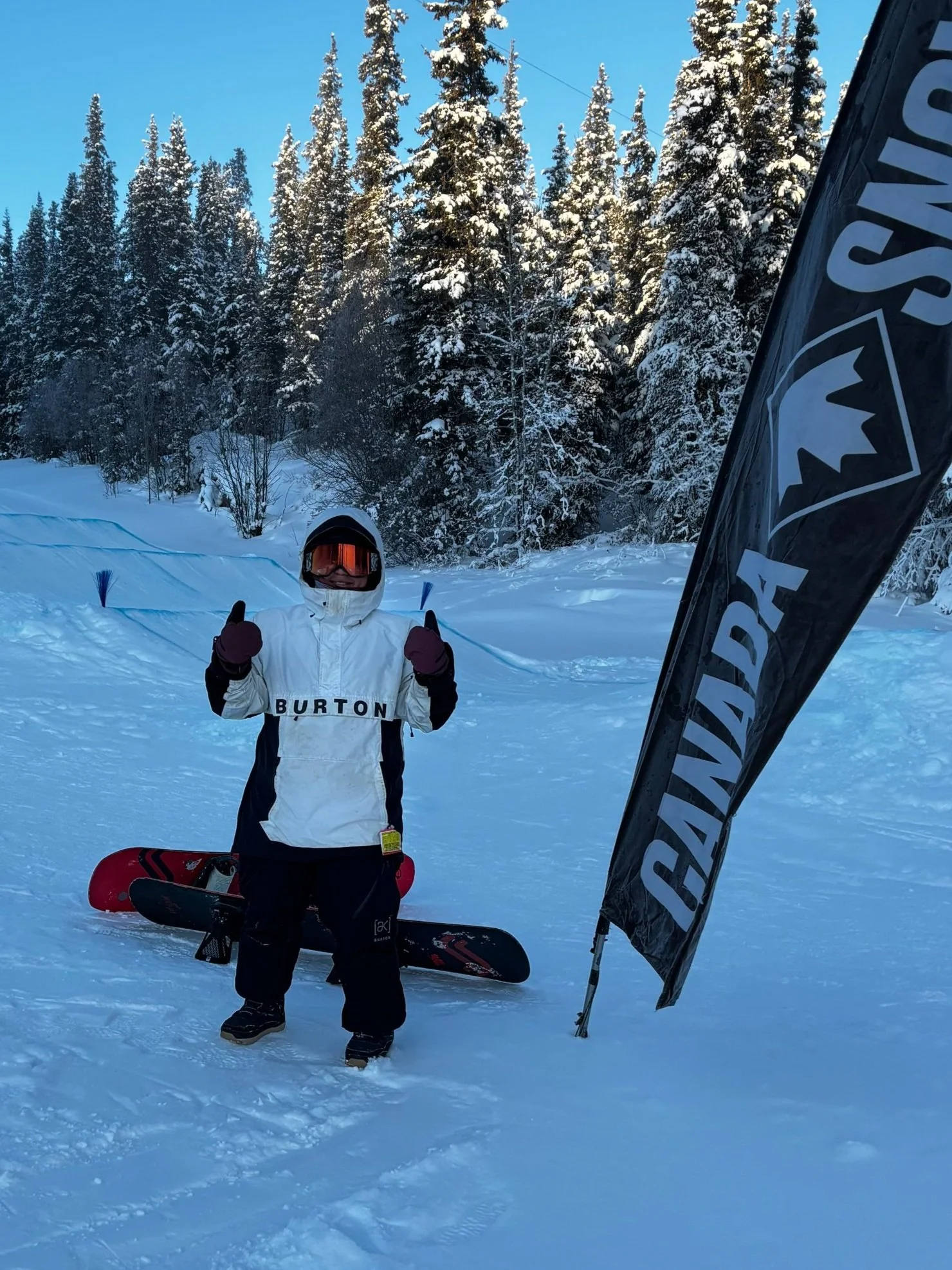 A person dressed in winter ski gear standing in a snowy landscape with snow-covered trees in the background, giving a thumbs-up gesture. There is a snowboard on the ground and a black flag with the Burton brand logo.