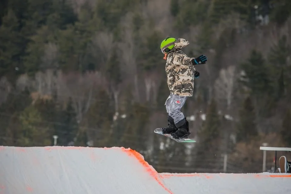 A young person in winter clothing, including a camouflage jacket, bright green helmet, and gloves, is mid-air on a snowboard in a snowy outdoor skate park with a snow bank and trees in the background.