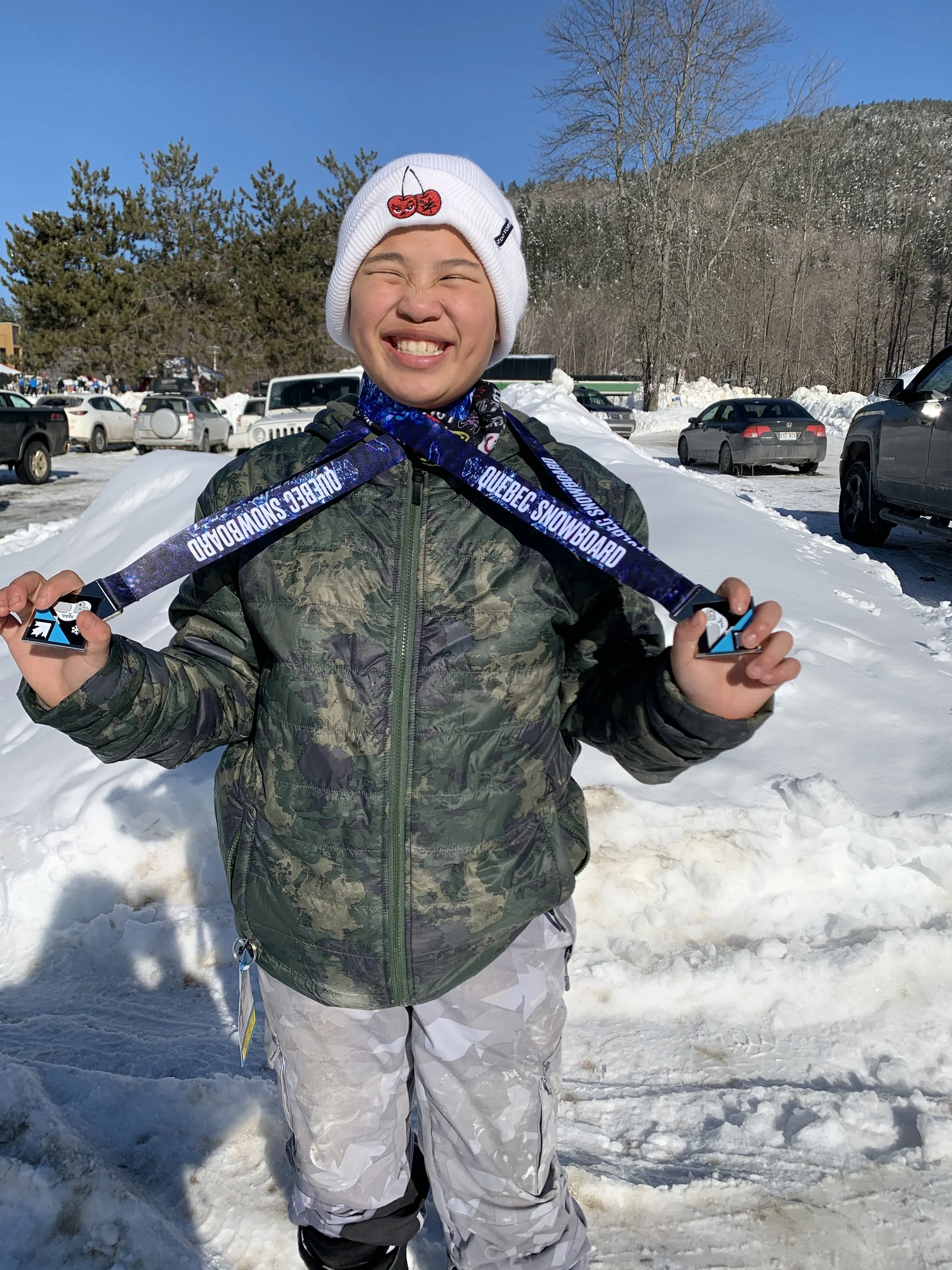 A young person smiling and holding a snowboard medal in a snowy outdoor parking area.