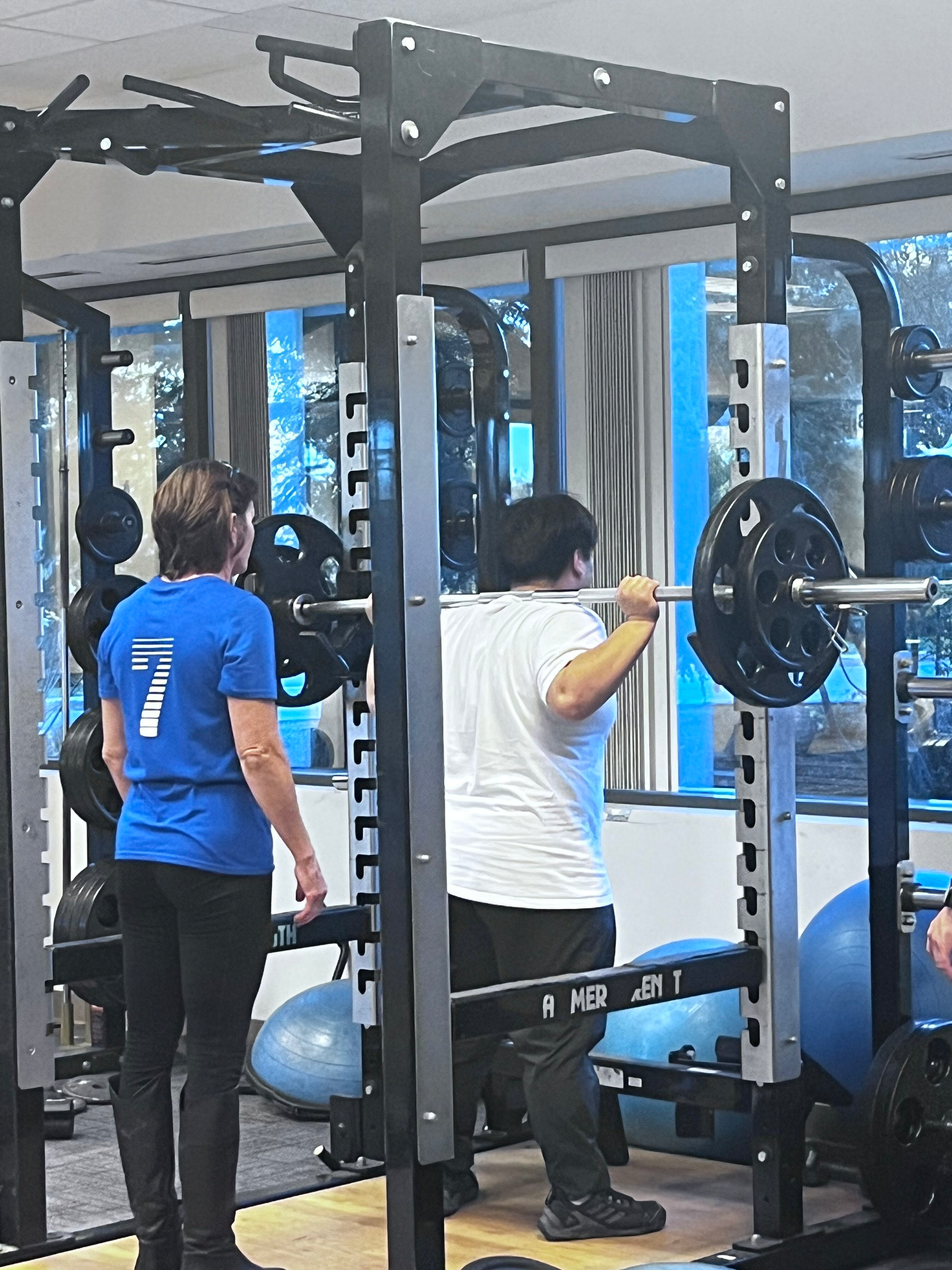 A man in a white t-shirt is performing a barbell squat with weights, while a woman in a blue shirt observes him at the gym. The gym has large windows and various exercise equipment.