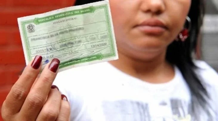 Mulher segurando carteira de identidade brasileira, com parte do rosto visível, esmalte vermelho nas unhas e brincos de flor.
