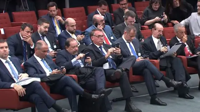 Homens de terno sentado em poltronas vermelhas em uma conferência, alguns usando celulares.