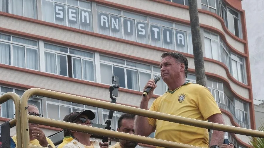 Homem falando ao microfone durante protesto, com pessoas ao redor, em frente a prédio, com cartaz no edifício que diz "SEM ANISTIA".