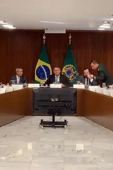Reunião oficial com homens ao redor de uma mesa, com bandeiras do Brasil ao fundo.