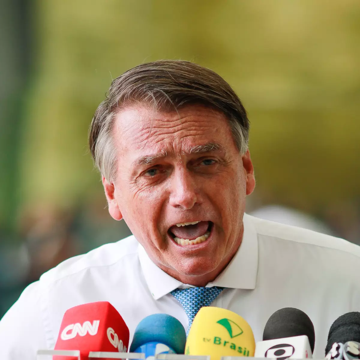 Homem falando durante uma entrevista ou coletiva, rodeado por microfones de várias emissoras de TV.