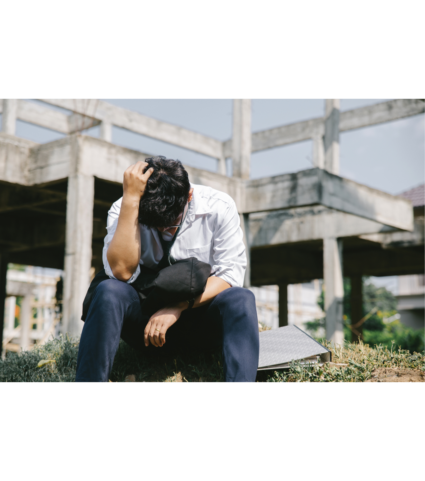 Homem sentado no chão, parece estar preocupado ou estressado, com uma mão na cabeça e outro segurando um chapéu, ao lado de um laptop aberto, em um local externo com estrutura de construção em segundo plano.