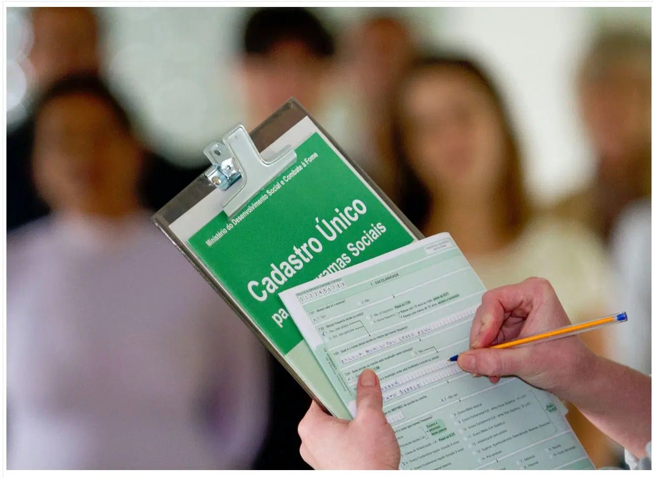 Pessoa preenchendo formulário de cadastro social com grupo de pessoas ao fundo, focado na mão com caneta e documentos.