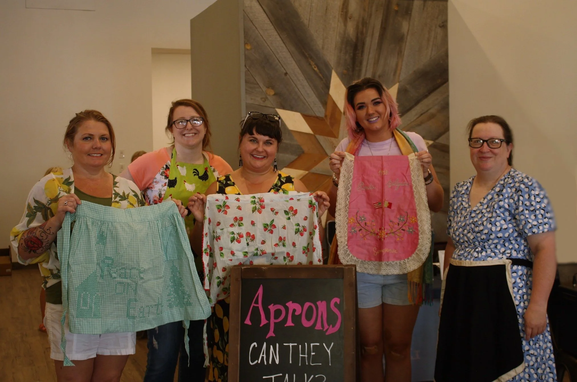 Group of women holding up historical aprons by a sign reading, "Aprons, can they talk?"