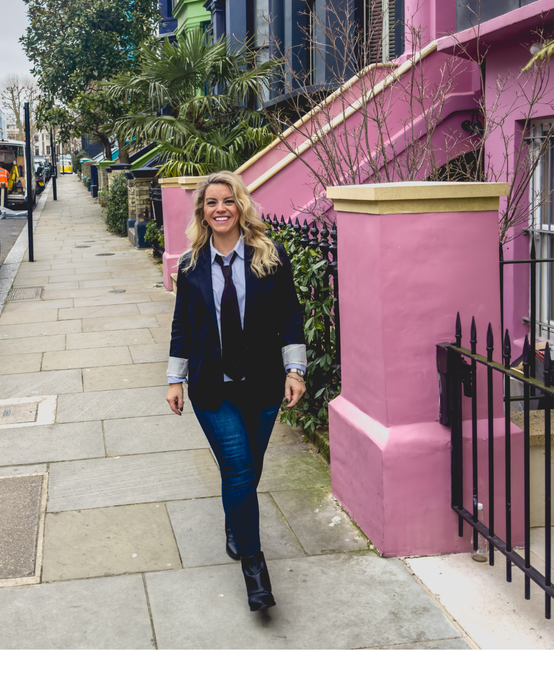 A woman smiling and walking on a city sidewalk in front of a pink building with a yellow accent, green plants, and a black iron fence.