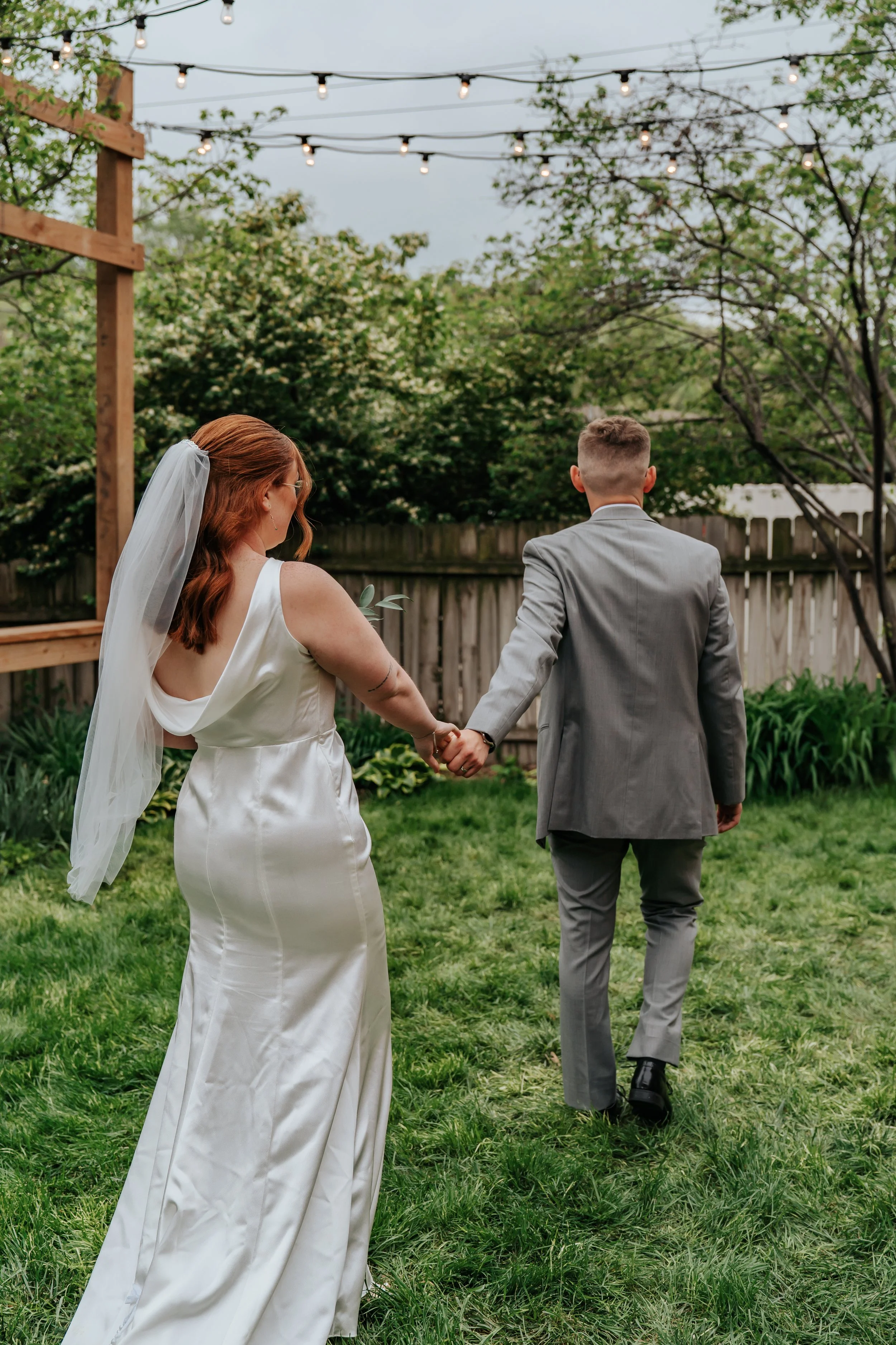 Bride and groom holding hands, walking in a backyard with string lights overhead and lush greenery, during a wedding celebration.
