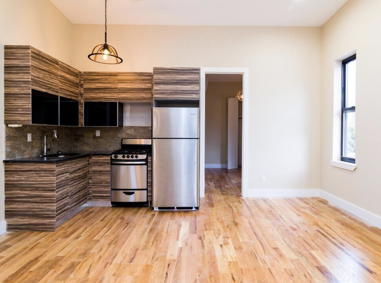 Empty kitchen with wooden cabinets, stainless steel refrigerator, stove, black countertop, wooden flooring, a window, and an open doorway.
