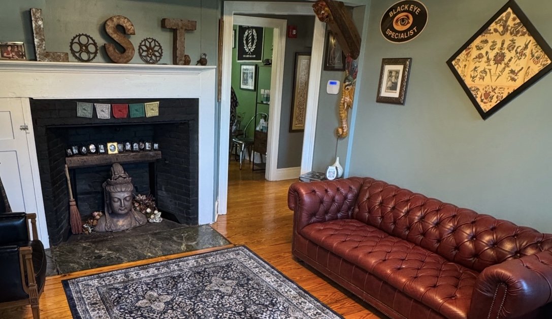 Living room with a brick fireplace, a red leather chesterfield sofa, a patterned area rug, wall decorations including a large eye sign, framed pictures, and a doorway leading to another room with green walls.
