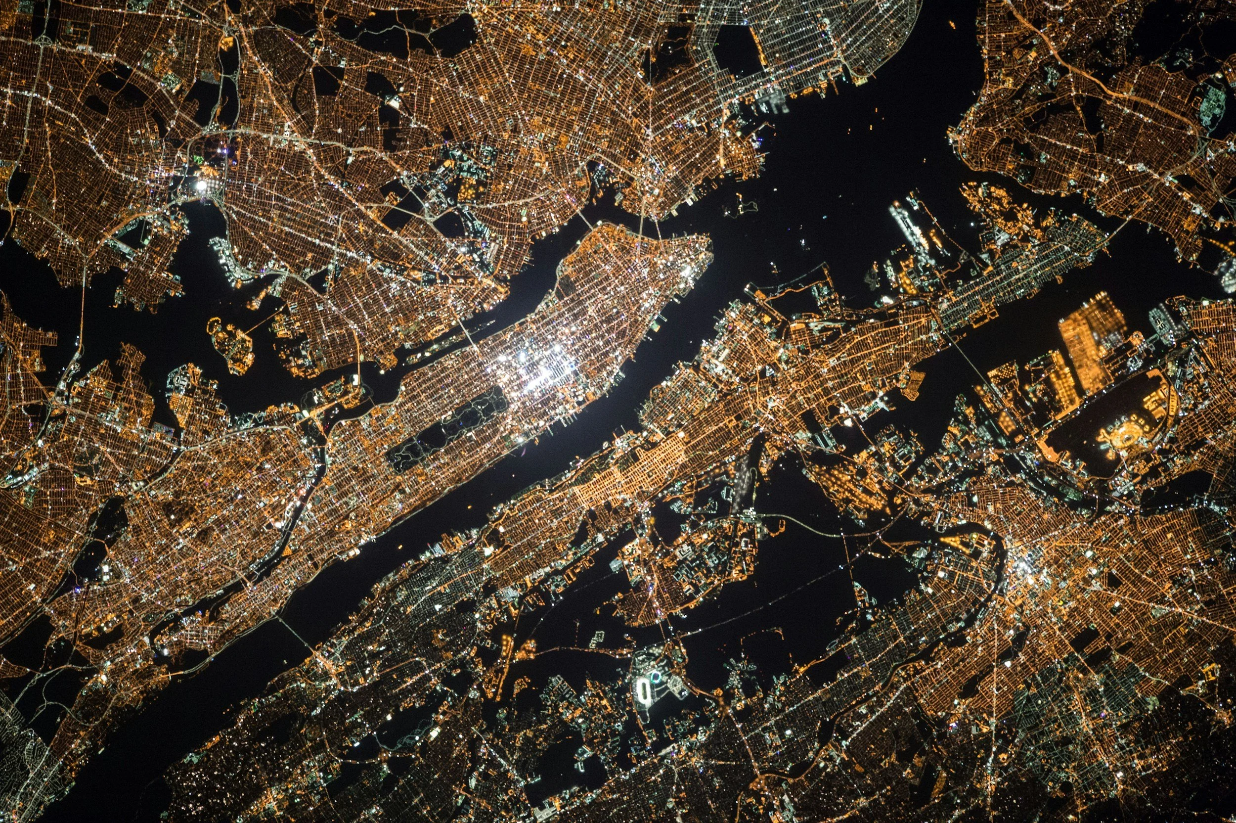 An aerial nighttime view of a city with illuminated streets and buildings. The city is surrounded by water with bridges connecting different areas.