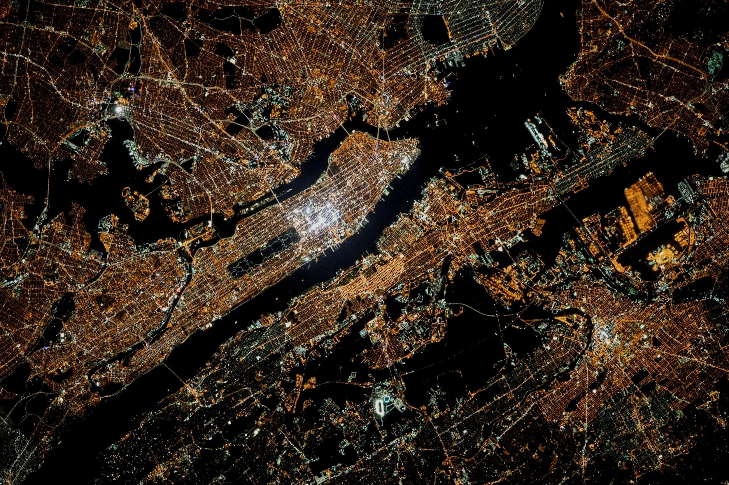 Aerial night view of Manhattan in New York City with illuminated streets, bridges, and water bodies.