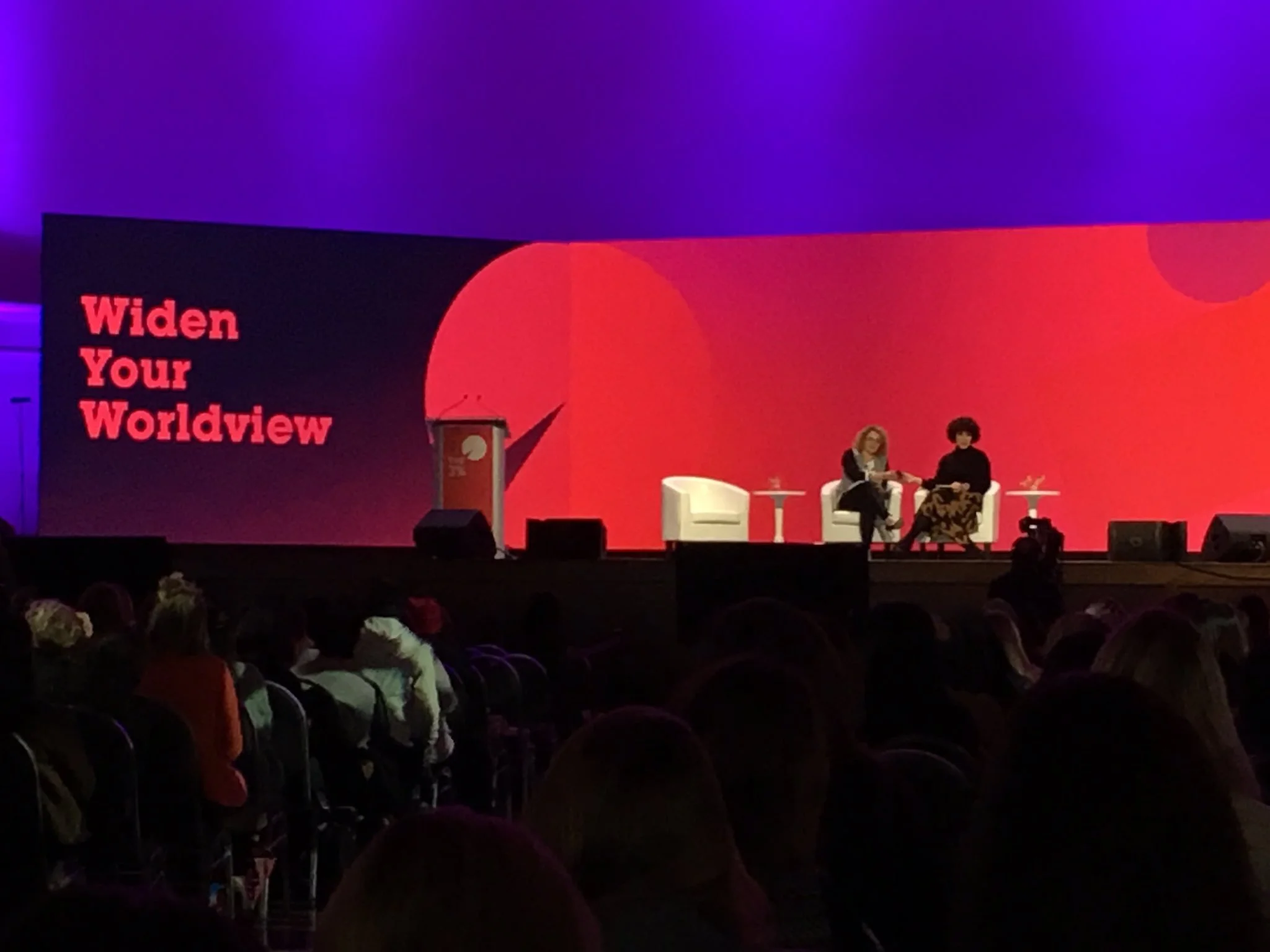 At an event stage, two women are sitting on white chairs engaged in conversation, with a large red and black screen behind them displaying the partial phrase "Widen Your Worldview." Audience members are visible in the foreground.
