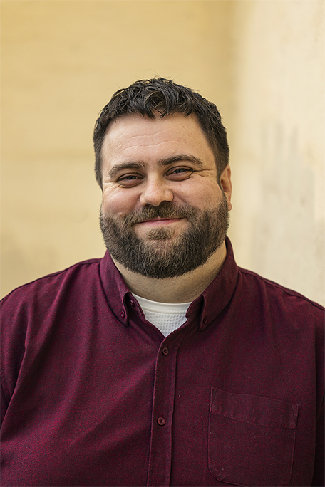 En man med mörkt, lockigt hår och skägg, klädd i en vinröd skjorta, står framför en ljus vägg.