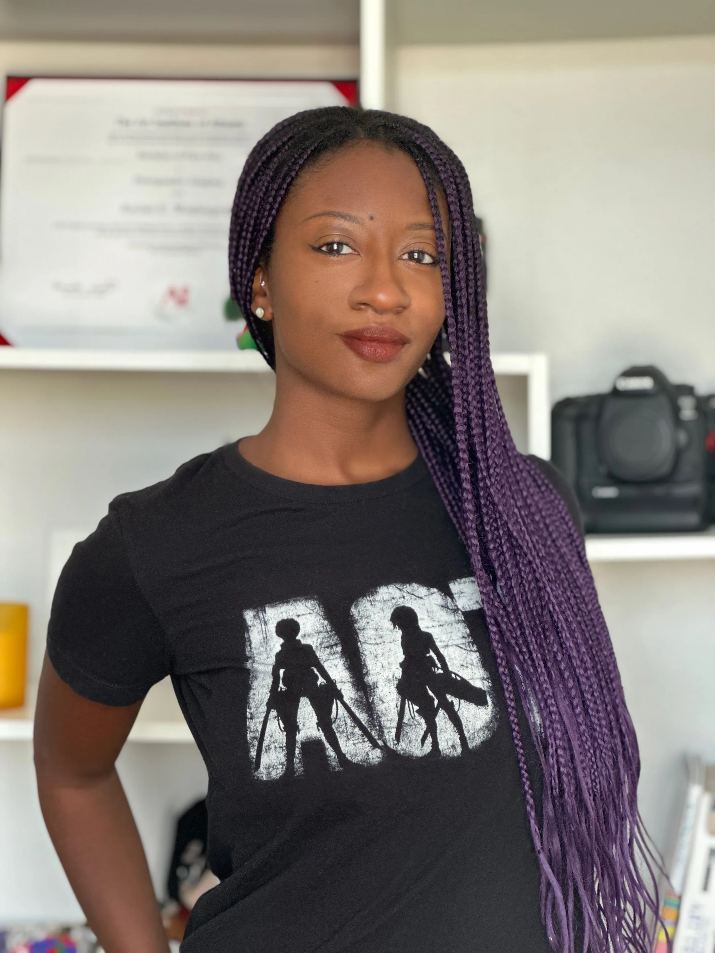 A young woman with purple braided hair, wearing a black T-shirt with a graphic of two people with backpacks, standing indoors near shelves and a camera in the background.