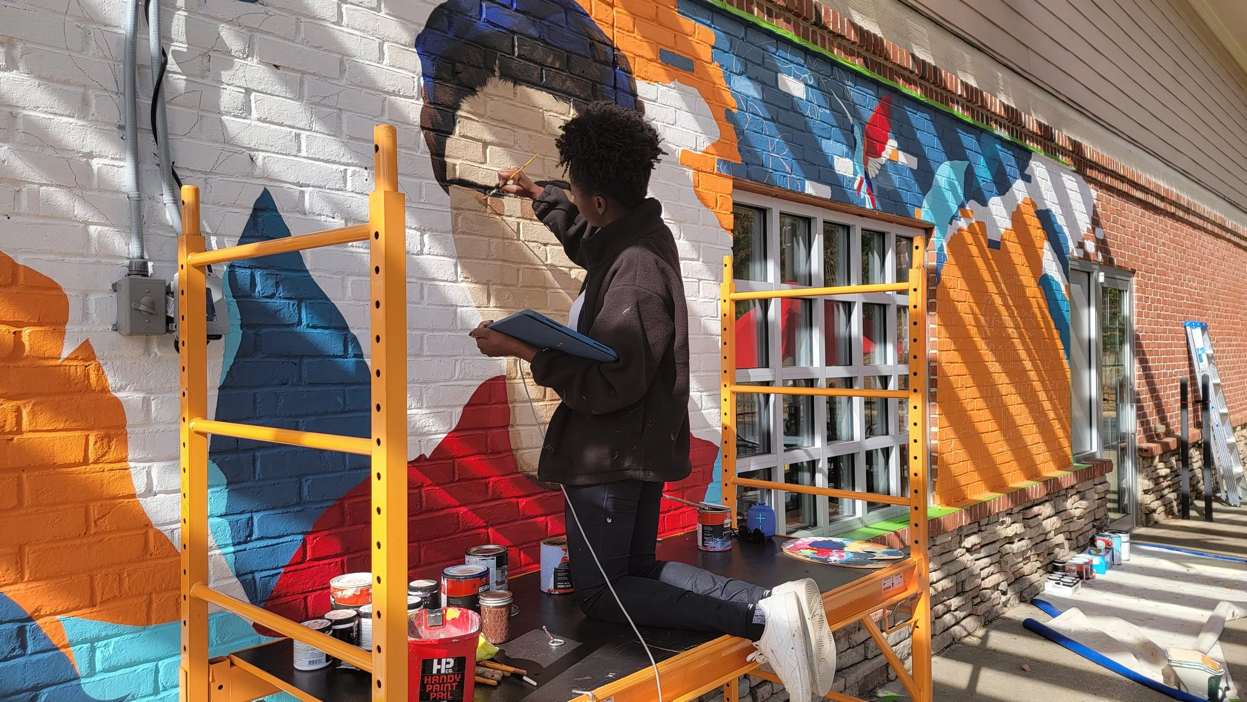 A young artist painting a colorful mural on a brick wall outside. She is kneeling on a black platform, holding a tablet, and using a small brush to add details to the mural.
