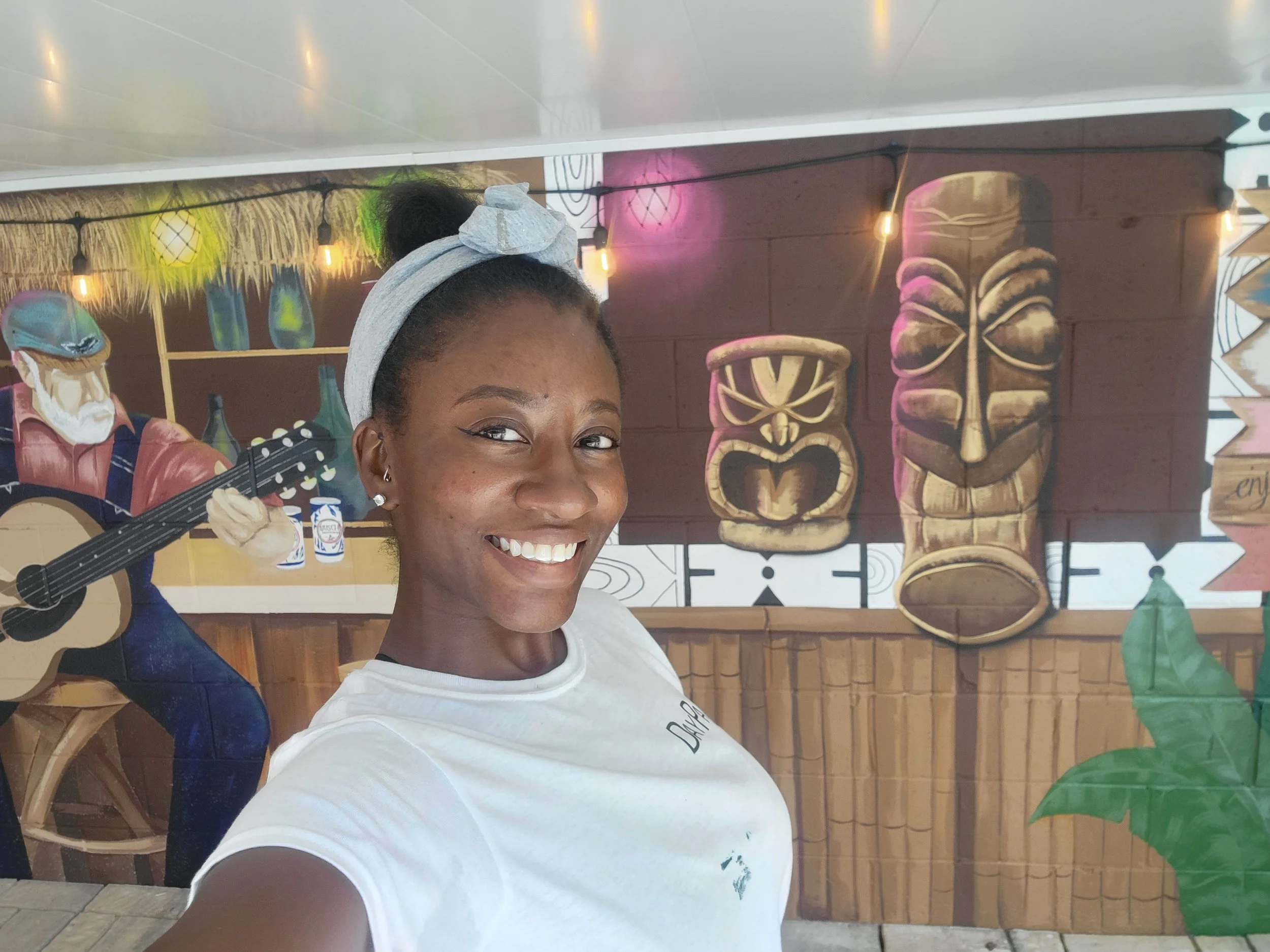 A woman with a gray headband and earrings smiling for a selfie in front of a colorful tiki bar mural, featuring tiki masks, a hula dancer, and string lights.
