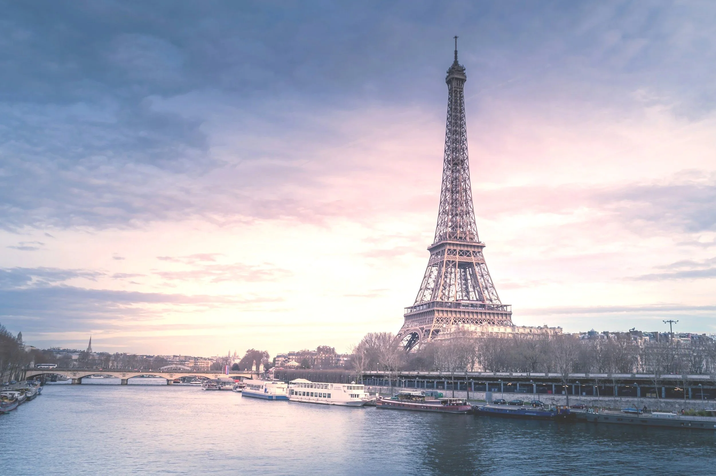 The Eiffel Tower in Paris, France, next to the river with boats docked along the shore during a sunset with partly cloudy skies.