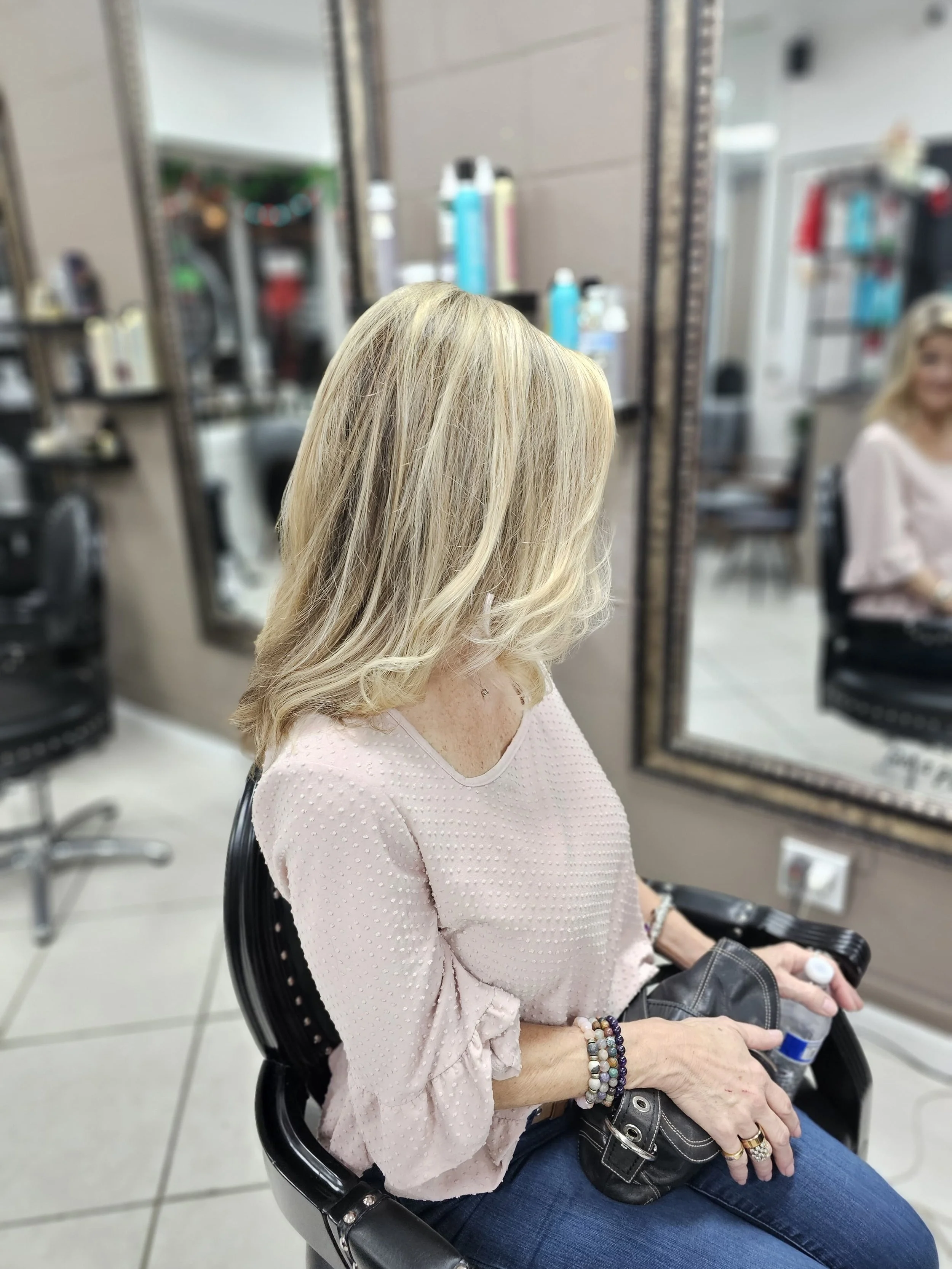 A woman with blonde, wavy hair sitting in a salon chair, reflected in a mirror. She is wearing a pink textured top, blue jeans, and bracelets, and holding a water bottle and a black purse.