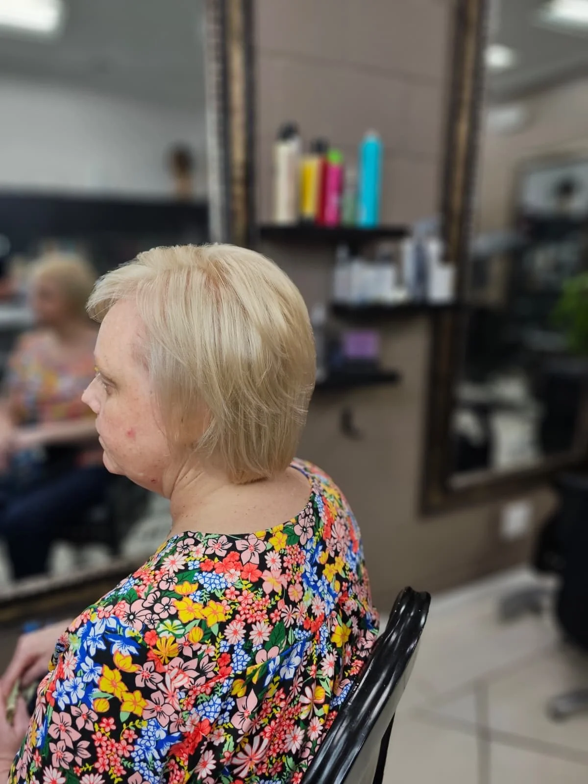 A woman with blonde hair sitting in a salon chair wearing a colorful floral top, with a reflection of herself in the mirror behind her.