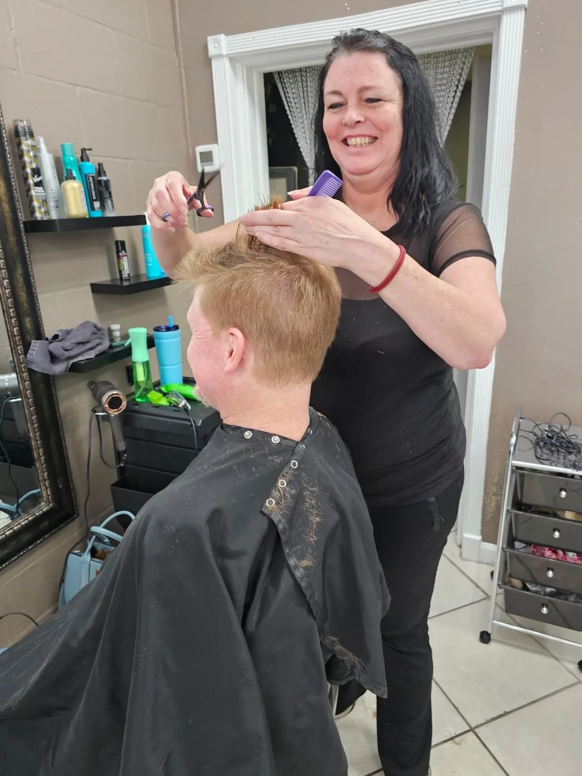 A woman cutting a man's hair at a salon, smiling and using scissors and a comb, with hair clippings visible on her black top and on the man's cape.