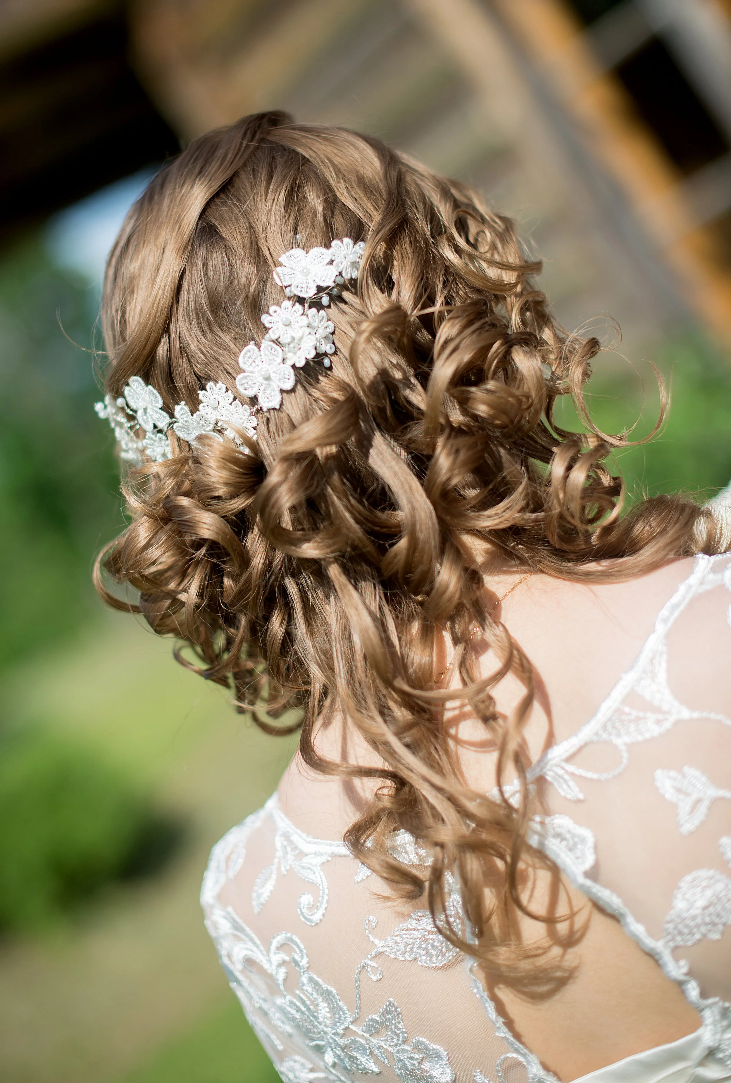 Back view of a woman with curly, light brown hair adorned with a white floral hair accessory, wearing a lace white dress.