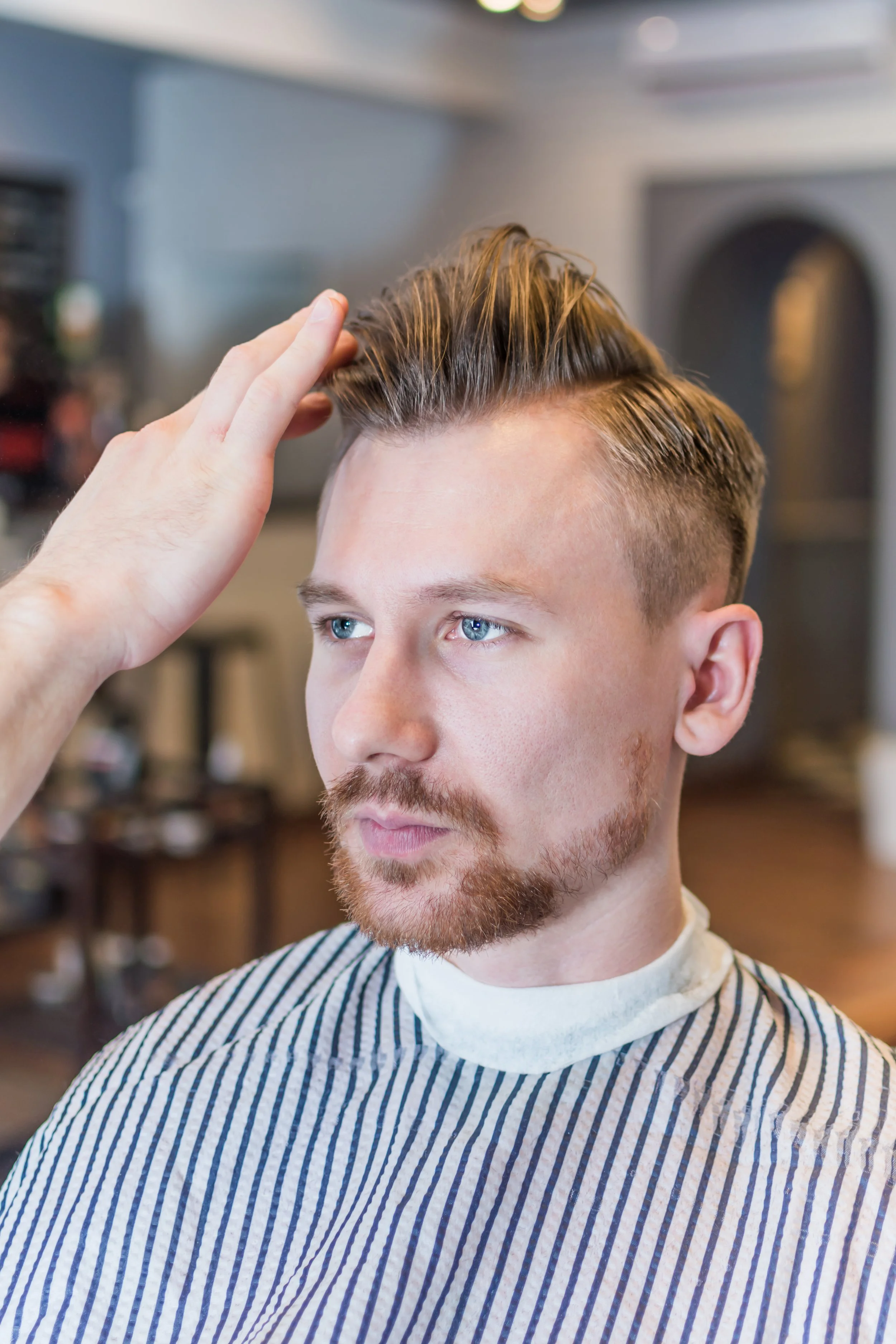 A man with blonde hair, blue eyes, and a beard getting a haircut at a barbershop, with a barber adjusting his hair.