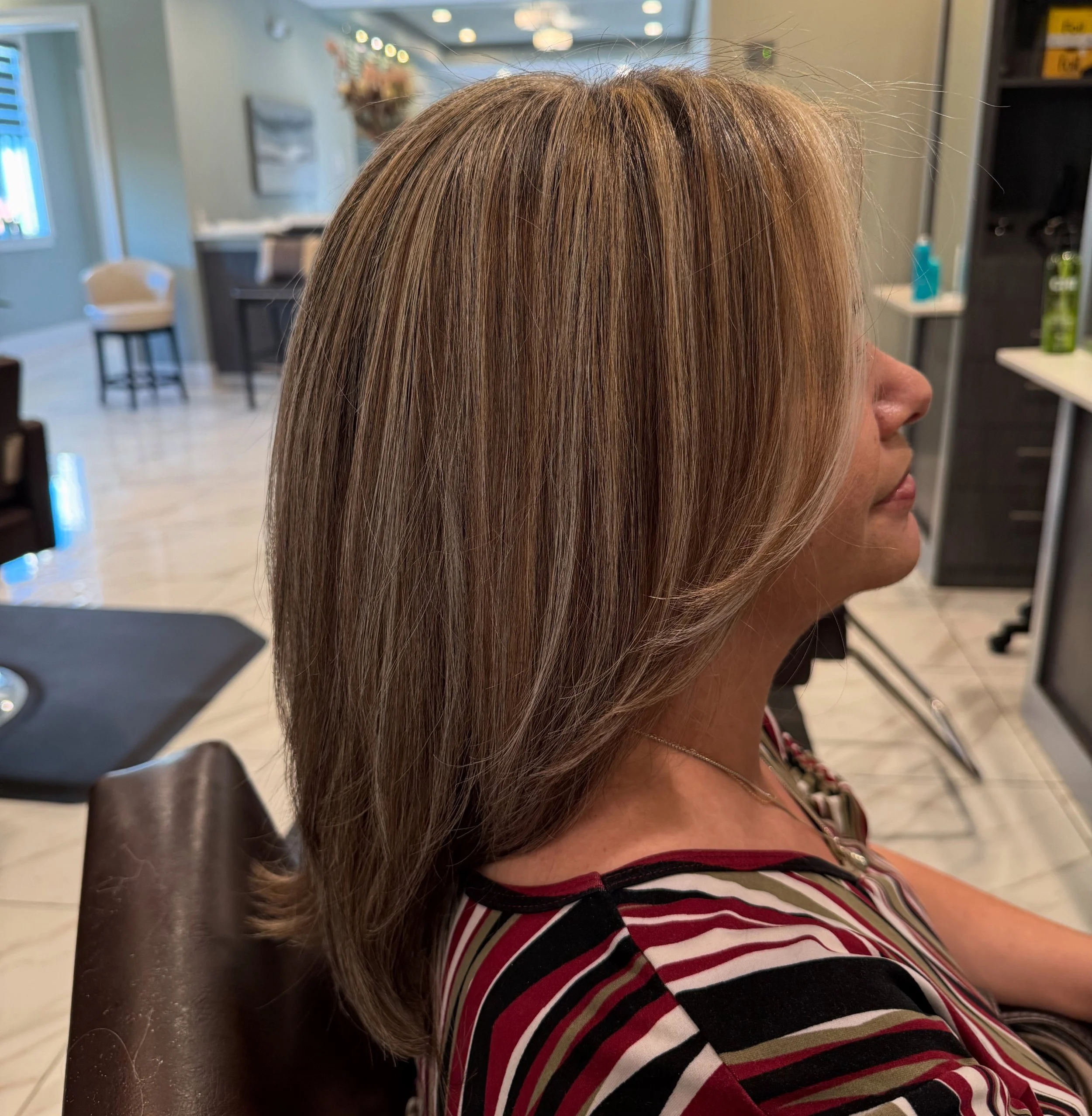 Side view of a woman with shoulder-length, straight, blonde hair sitting in a salon chair inside a hair salon.