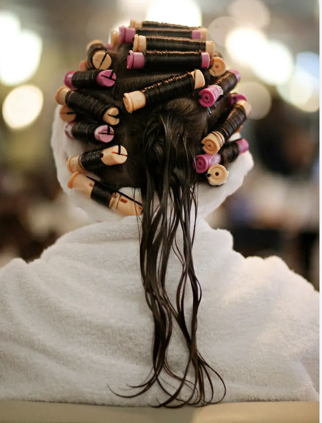 Person with dark hair in rollers, draped in a white towel, in a salon setting.